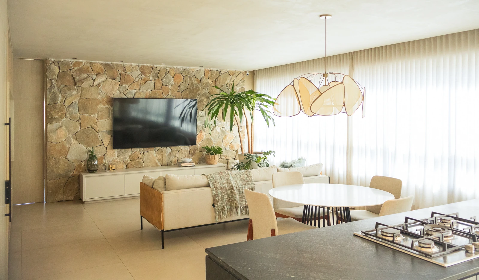 Sala de estar com parede de pedra natural, TV embutida, mesa de jantar redonda e ilha de cozinha com cooktop em primeiro plano