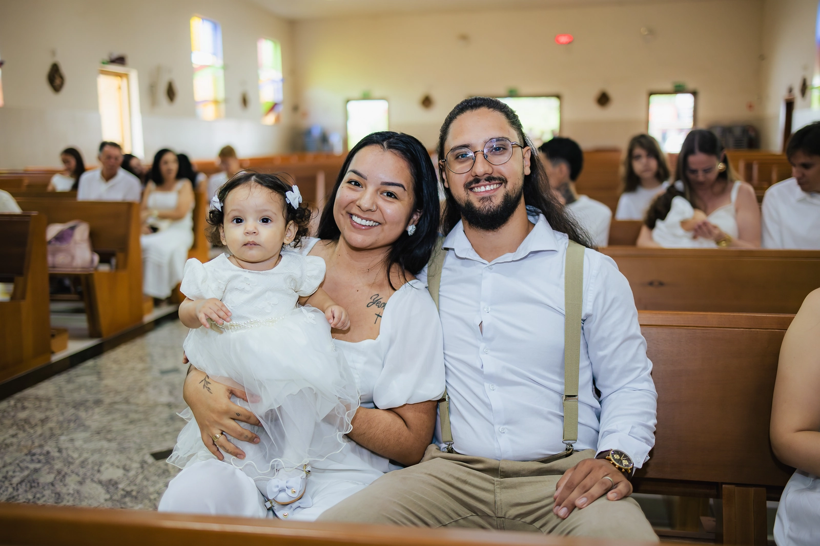 Pais sentados com a bebê no colo durante a cerimônia de batizado em Goiânia
