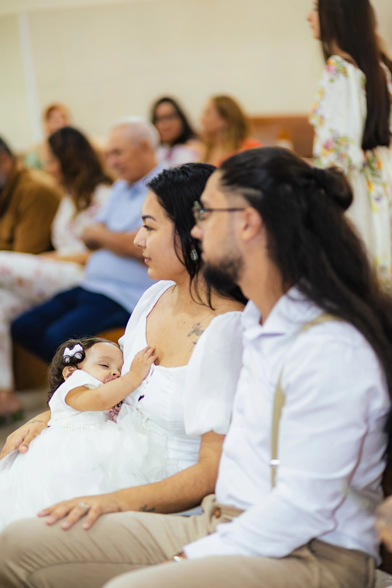 Pais com a bebê no colo acompanhando a cerimônia de batizado