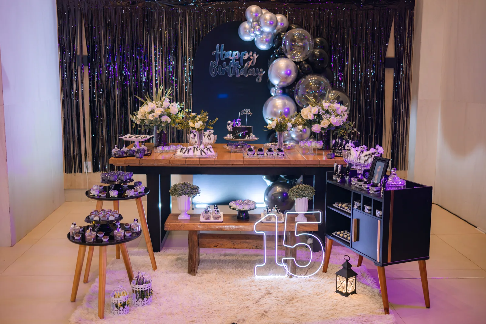 Mesa principal da festa de 15 anos da Sophya com painel Happy Birthday, balões prata e pretos e letreiro neon com o número 15