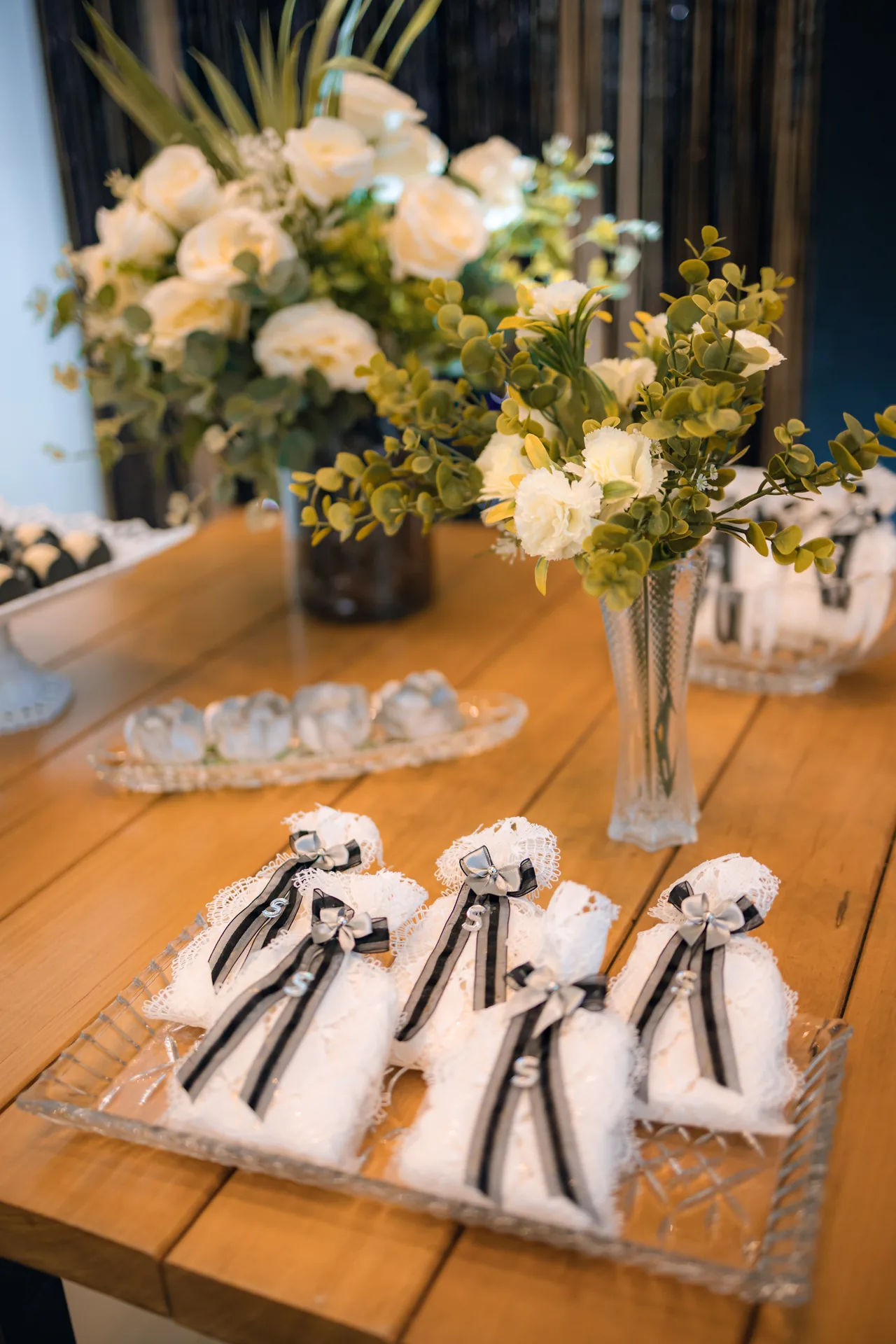 Doces embrulhados em tule branco com laço preto numa bandeja de cristal, com flores brancas ao fundo