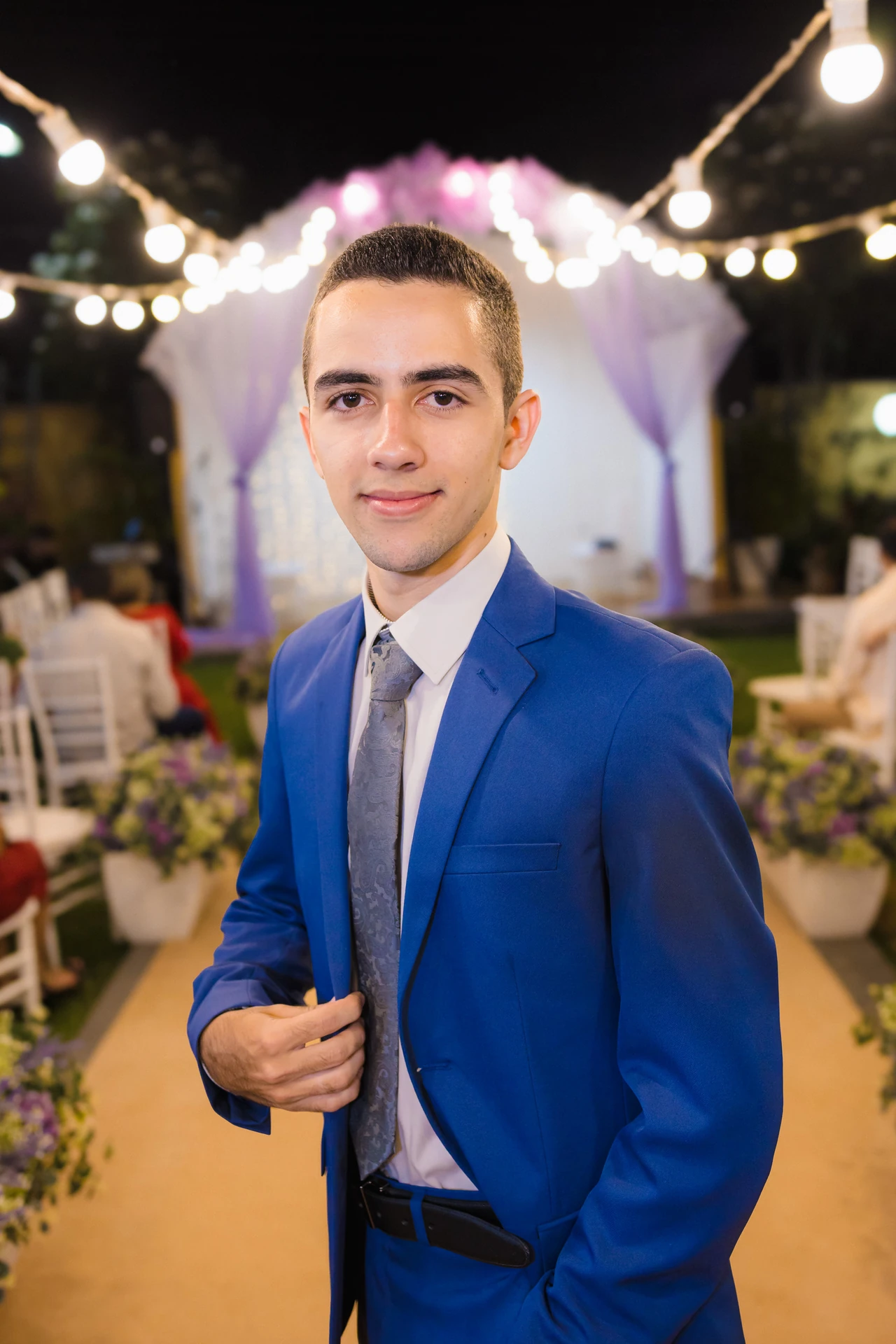 Retrato do noivo de terno azul e gravata cinza, sorrindo, no corredor da cerimônia iluminado por varal de luzes