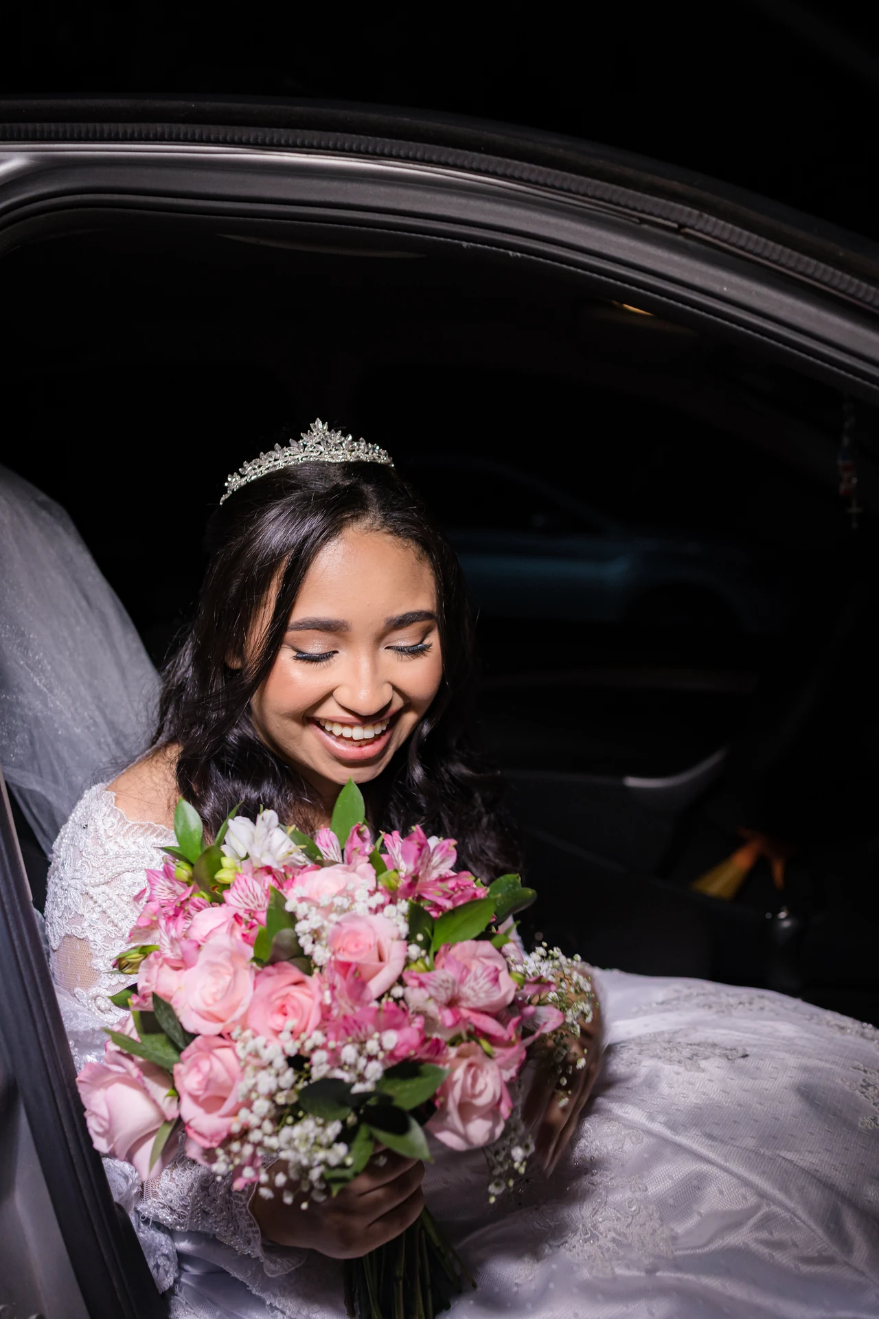 Noiva de tiara e vestido de renda sorrindo para o buquê de rosas e astromélias rosa, sentada no carro, à noite