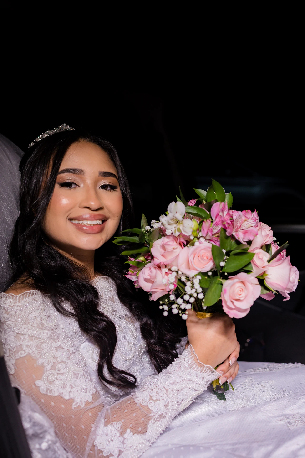 Noiva de véu e tiara sorrindo para a câmera com o buquê nas mãos, ainda dentro do carro, à noite
