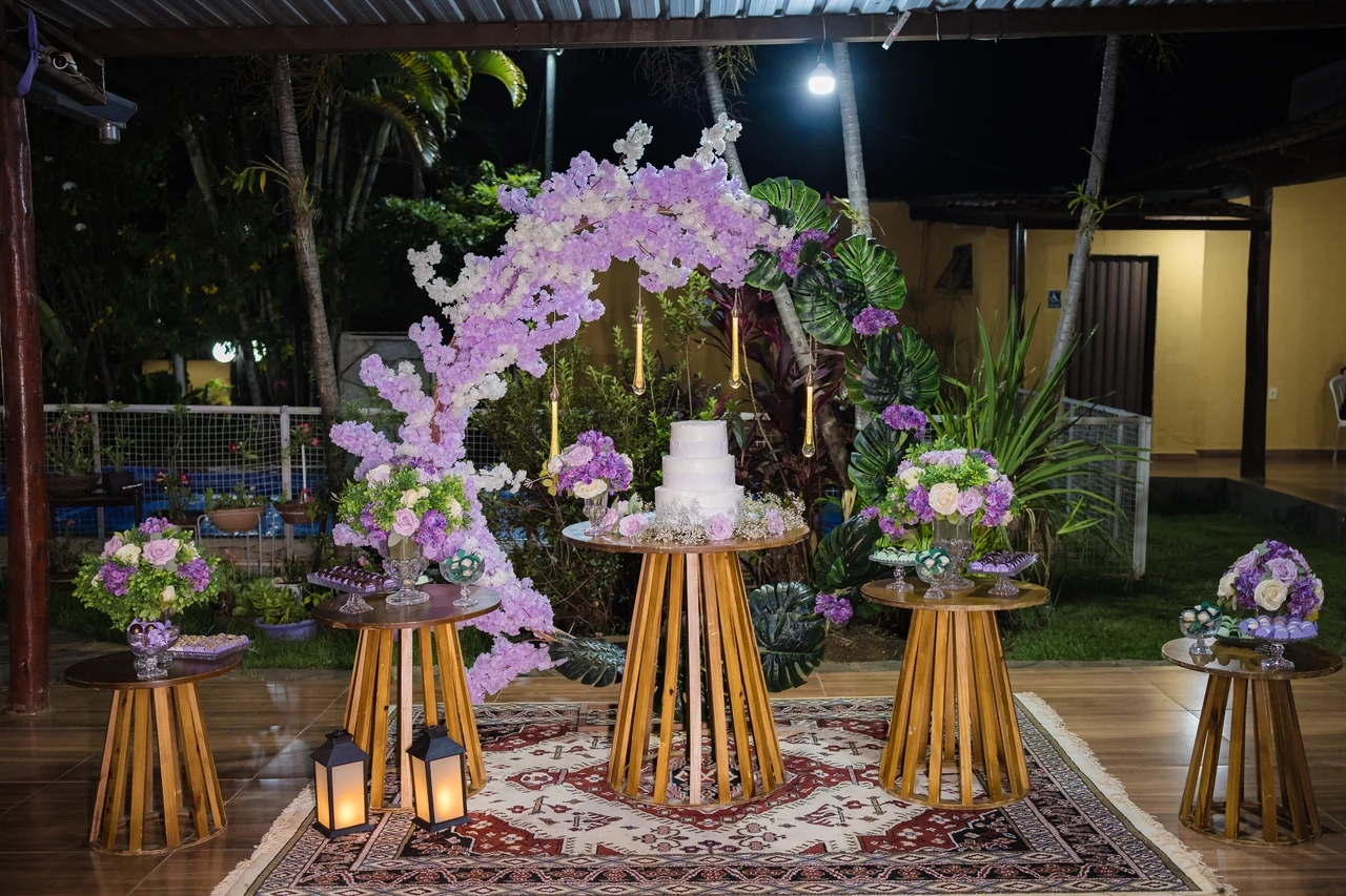 Mesa do bolo com arco de flores lilás, mesas de madeira com arranjos e tapete estampado, à noite