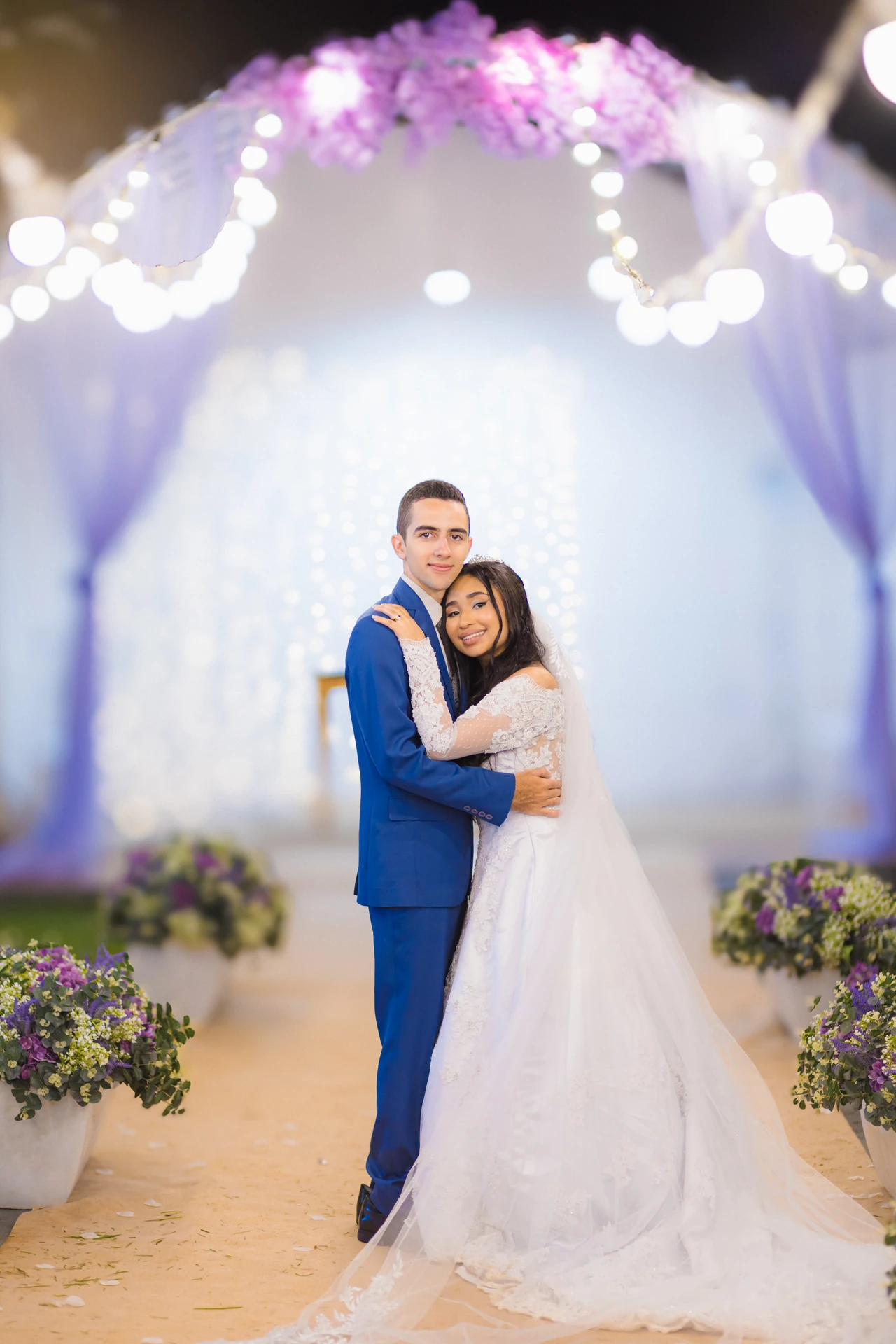 Casal abraçado e sorrindo para a câmera sob o arco de flores lilás e luzes, com o corredor de vasos ao fundo