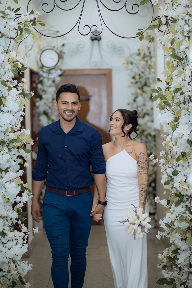 José e Alice sorrindo, de mãos dadas, sob um arco de flores brancas no Cartório Silva, em Goiânia