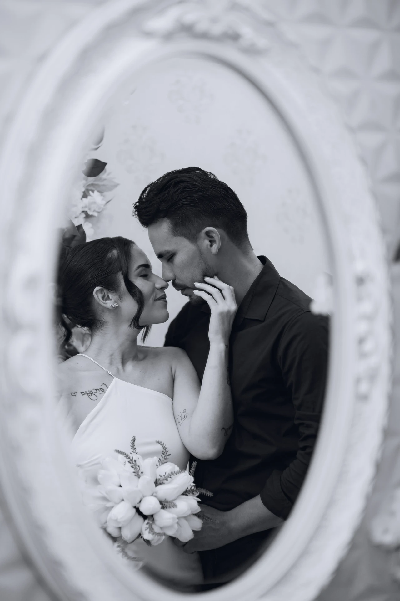 Casal em preto e branco enquadrado por um espelho oval branco, quase se beijando