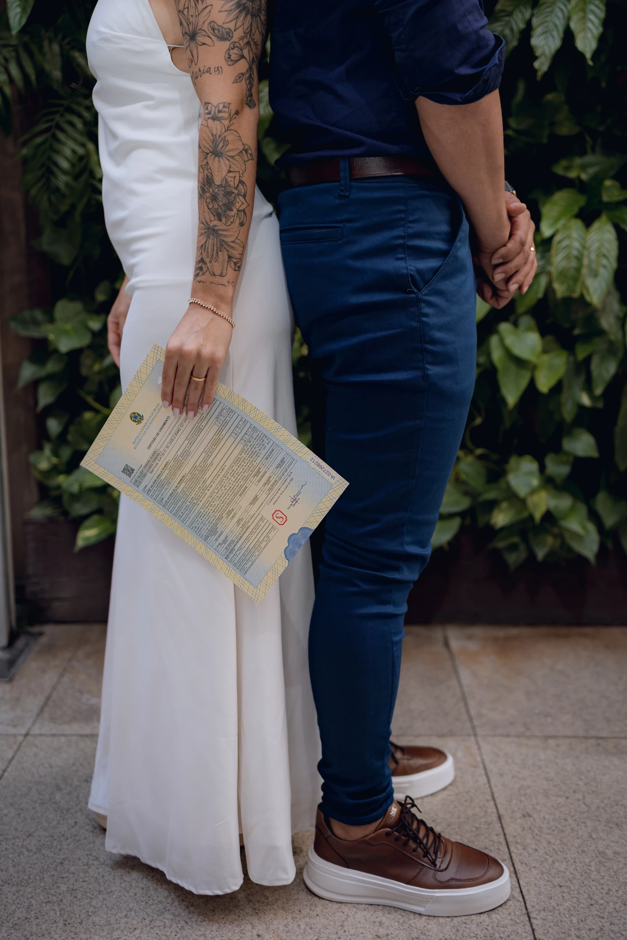 Noivos de costas diante da parede de folhagens, Alice segurando a certidão de casamento