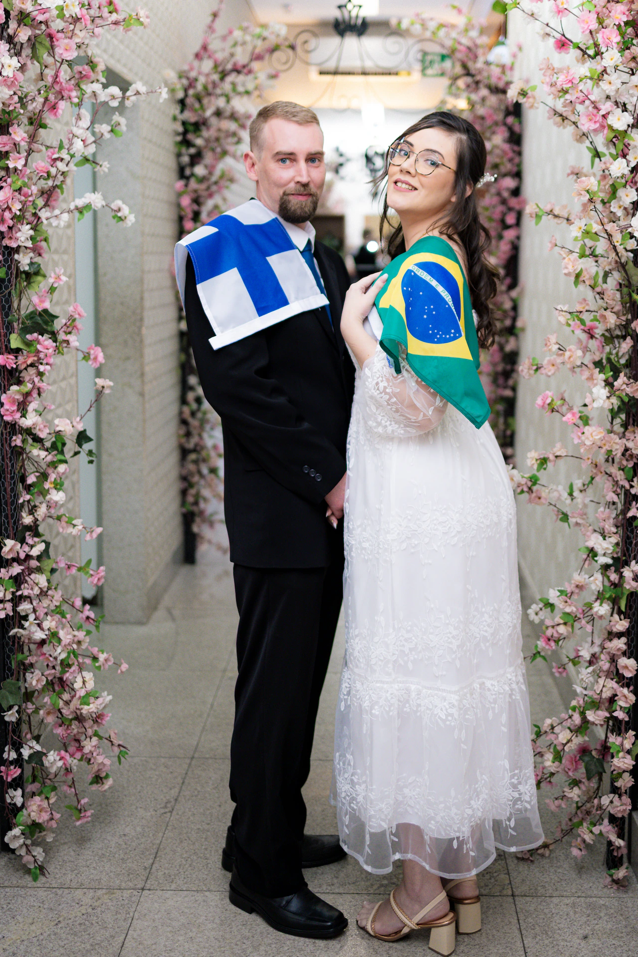 Os noivos sob o arco florido segurando as bandeiras da Finlândia e do Brasil, em casamento civil em Goiânia