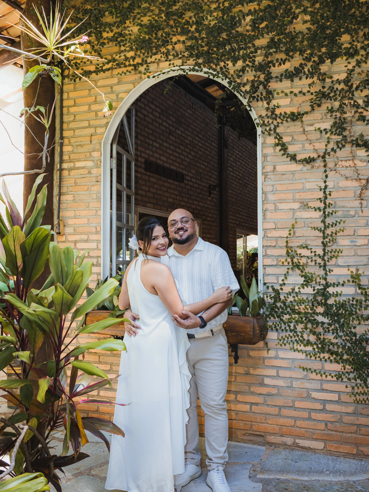 Jessica e Alex abraçados e sorrindo na entrada em arco de tijolo com plantas