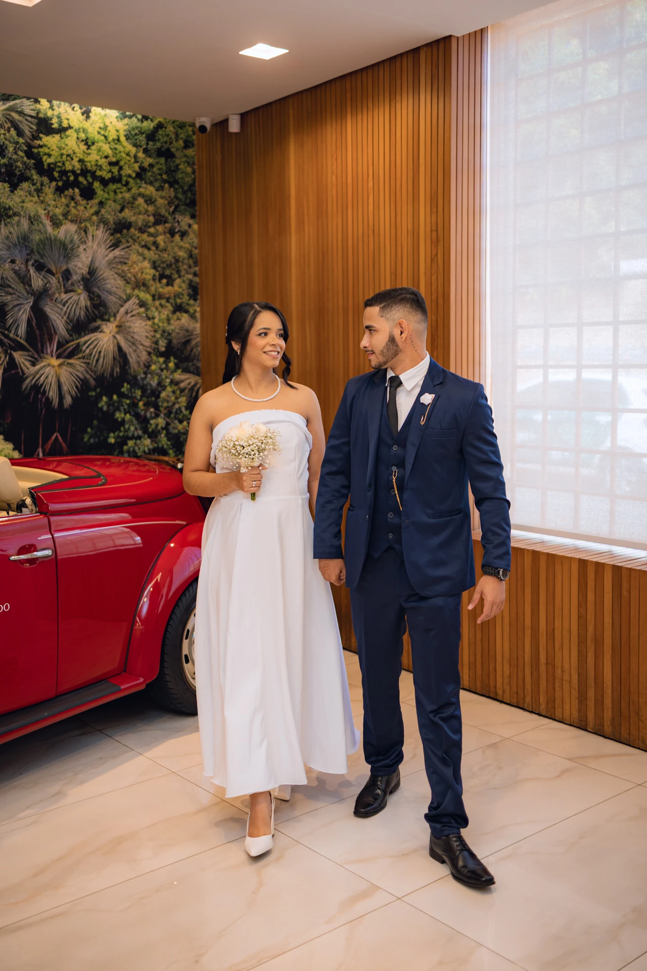 Jéssica e Thiago caminhando de mãos dadas ao lado do carro antigo vermelho