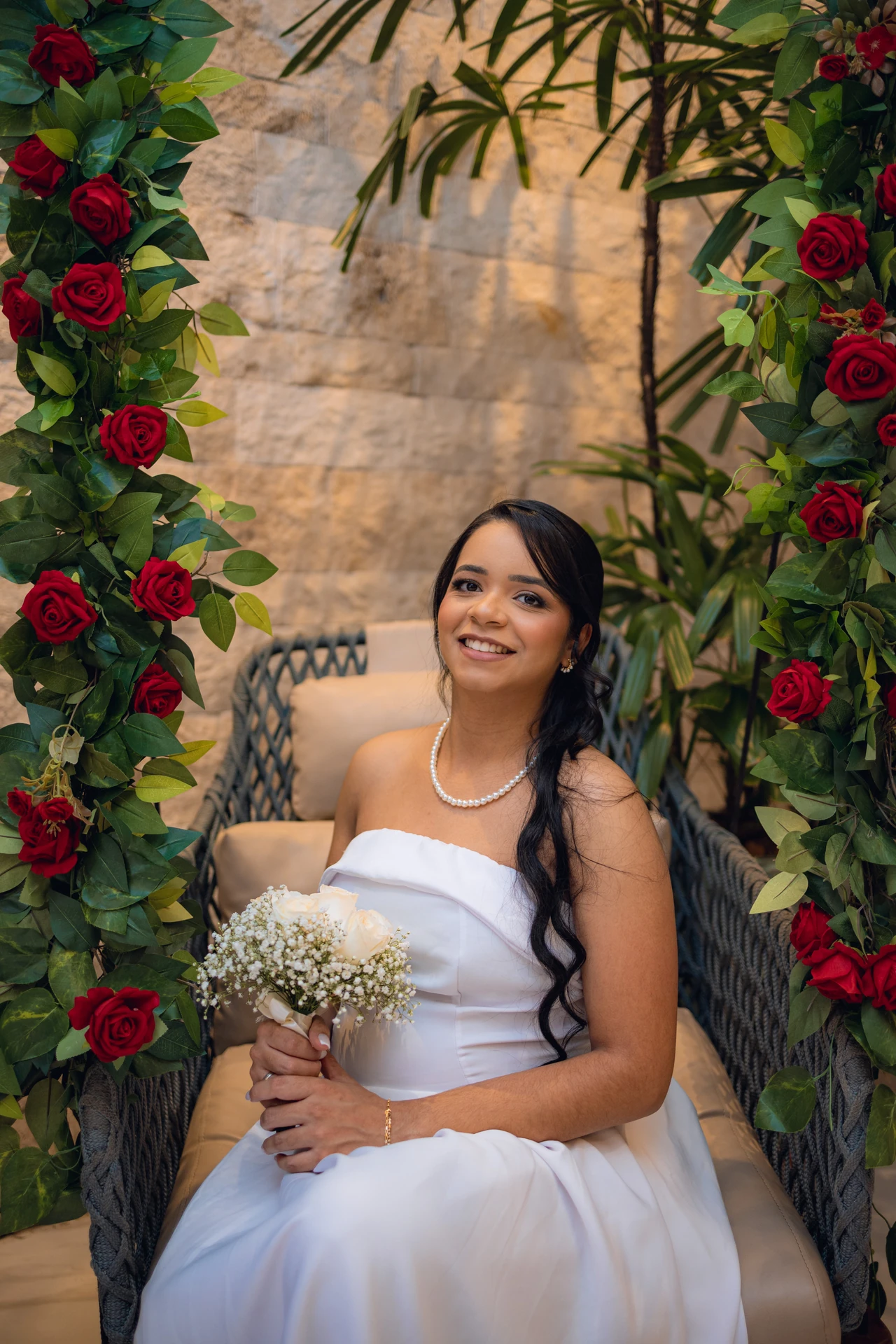 Jéssica sentada em uma poltrona de vime sob um arco de rosas vermelhas, segurando o buquê