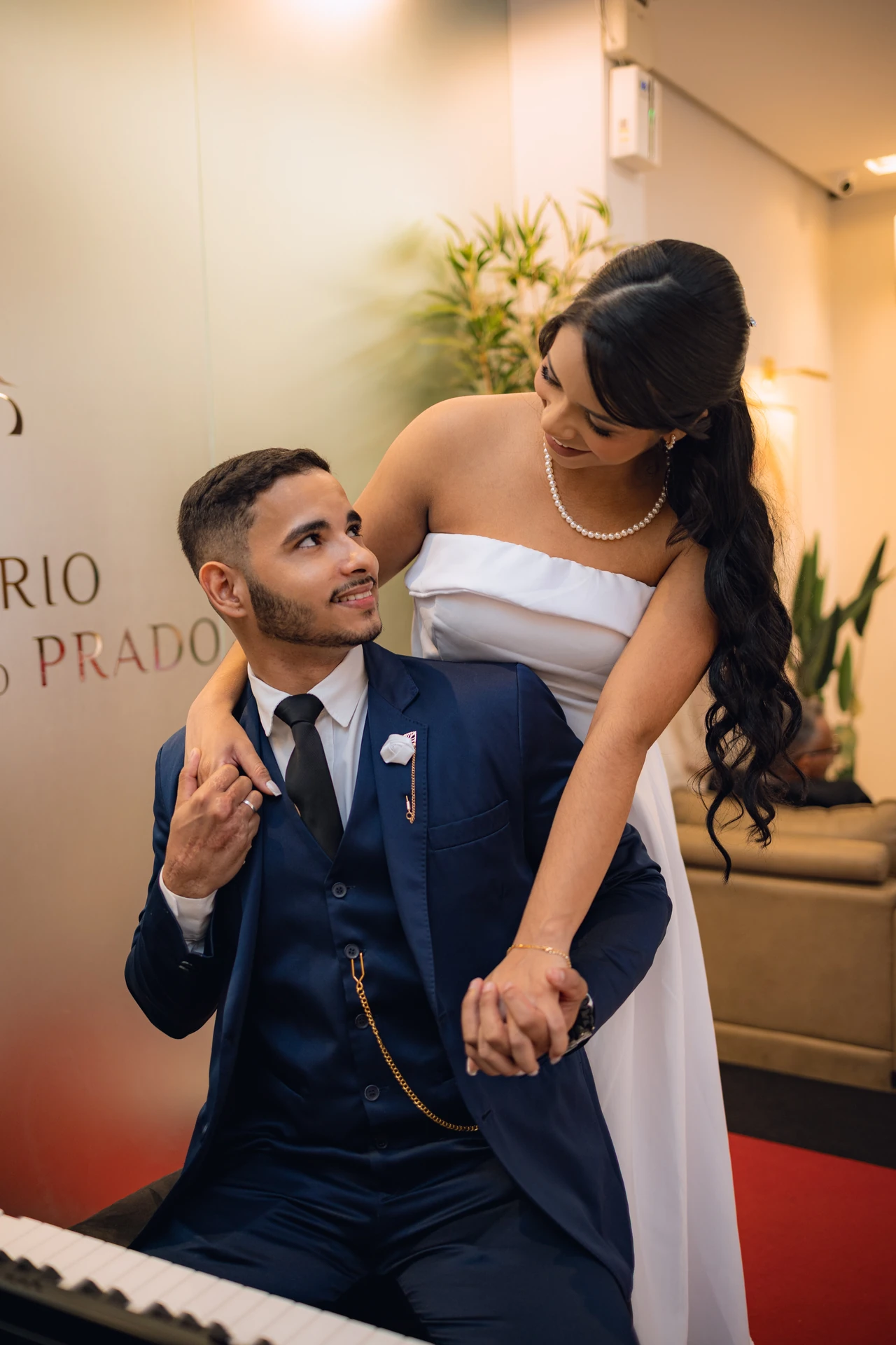 Jéssica abraçando Thiago por trás perto da porta de vidro com o logo do Cartório Antônio do Prado