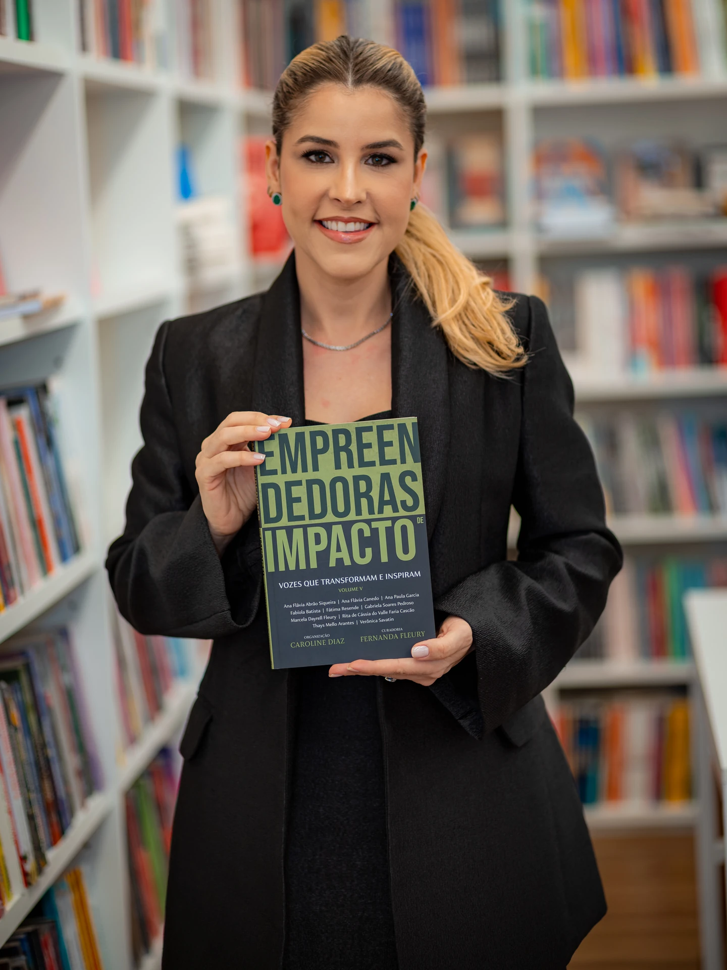 Autora de blazer preto sorrindo com o livro Empreendedoras de Impacto nas mãos, em frente às estantes da livraria