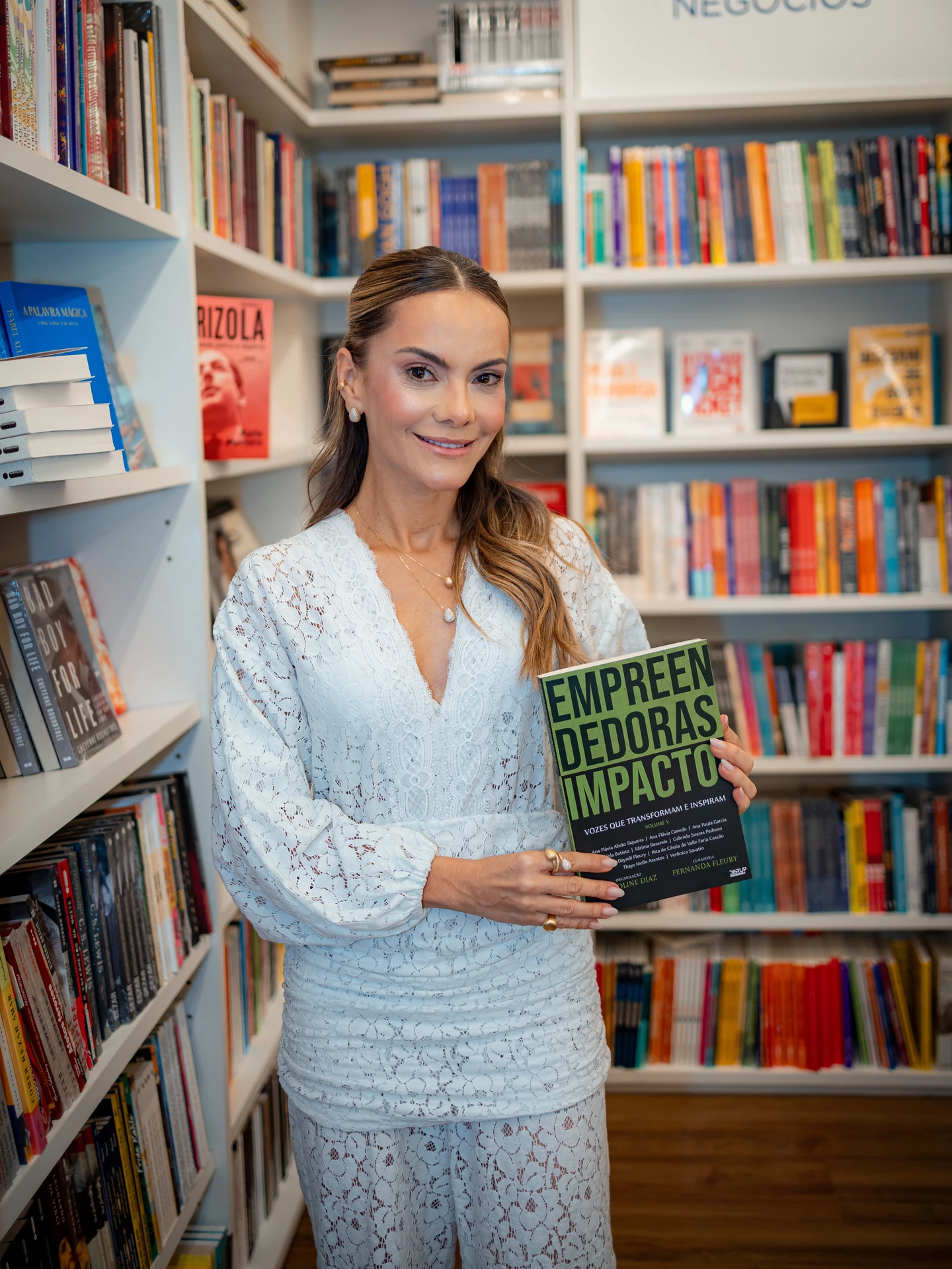 Autora de conjunto branco de renda com o livro Empreendedoras de Impacto nas mãos, entre as prateleiras da livraria