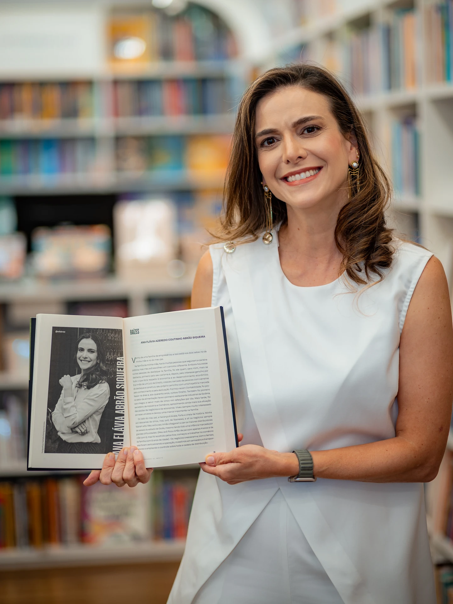 Autora de blusa branca sorrindo e mostrando a página do livro com o seu retrato