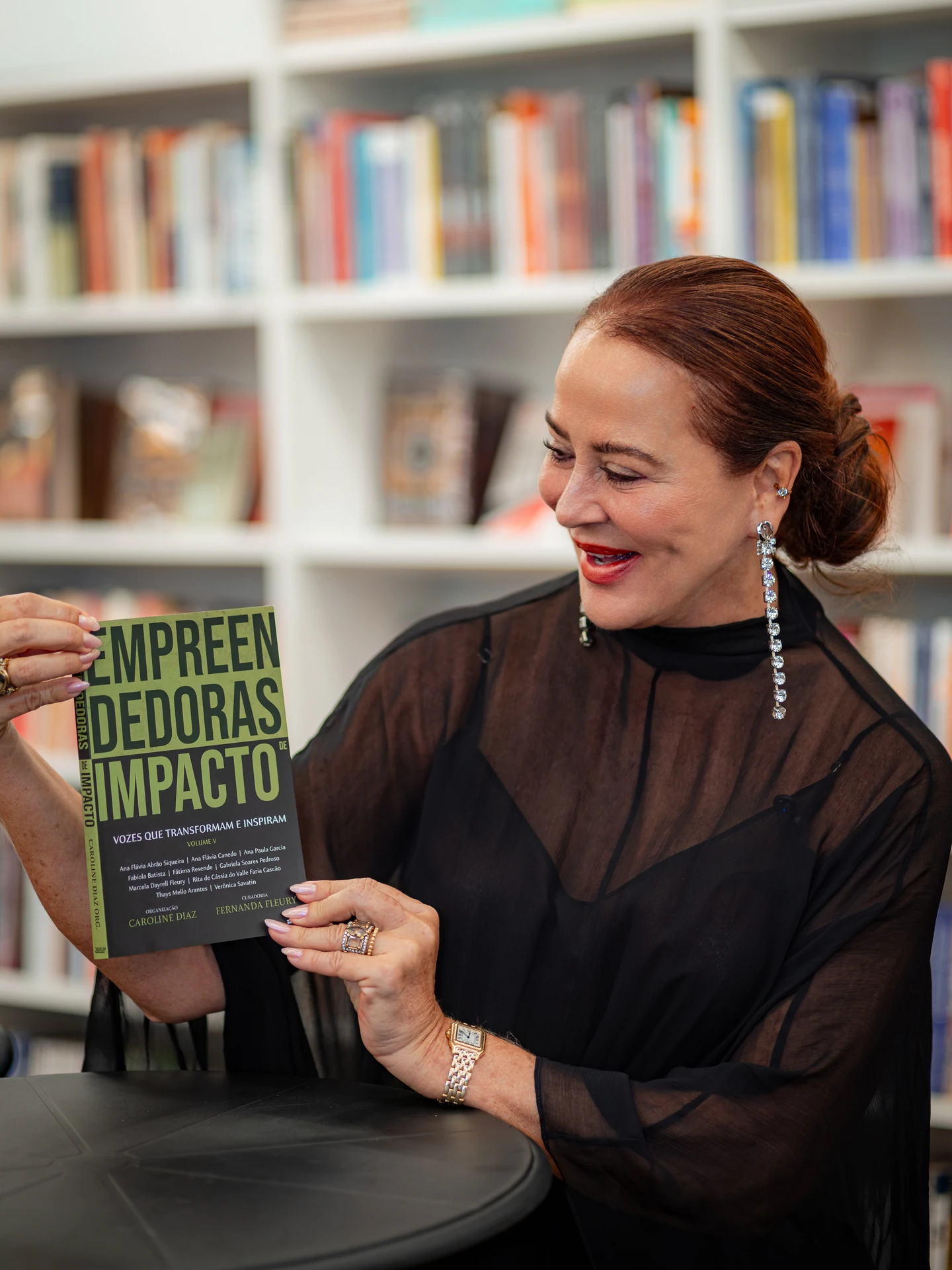 Autora de blusa preta transparente sentada à mesa, olhando para o livro Empreendedoras de Impacto que segura