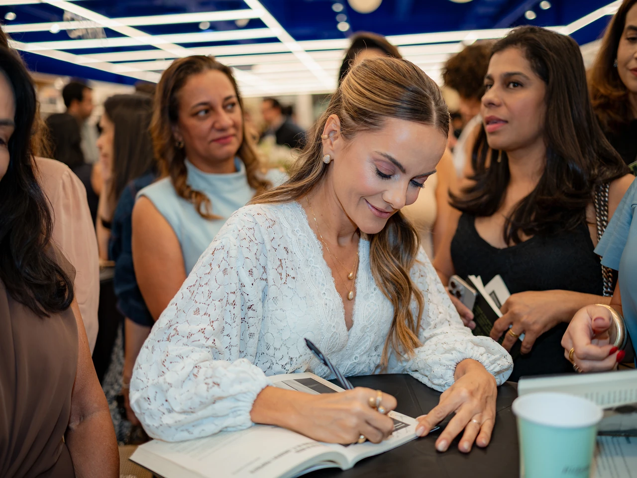 Autora de conjunto branco de renda autografando um livro na mesa, cercada de convidados na Livraria da Vila
