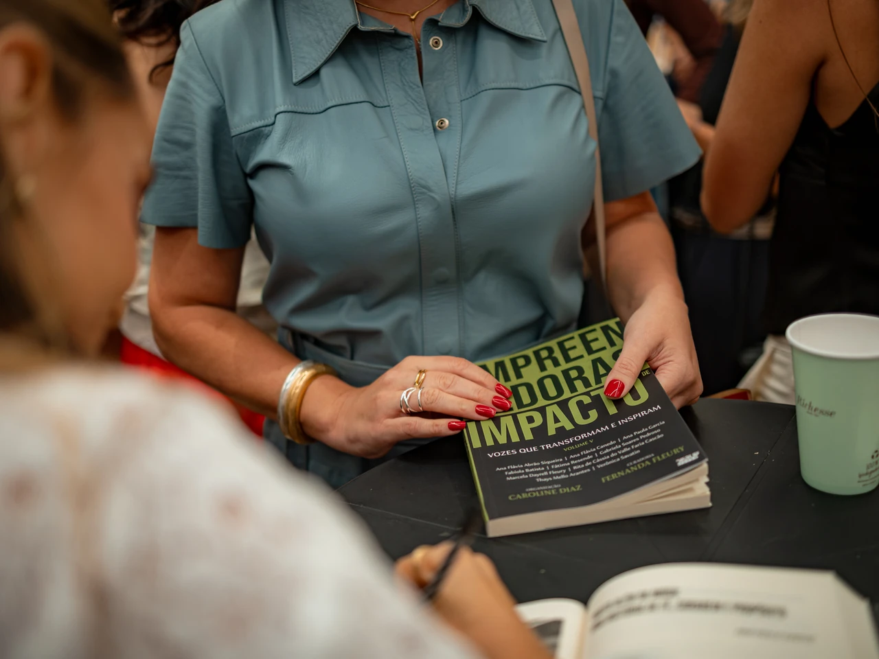 Convidada segurando um exemplar do livro na mesa de autógrafos, com outras pessoas ao redor