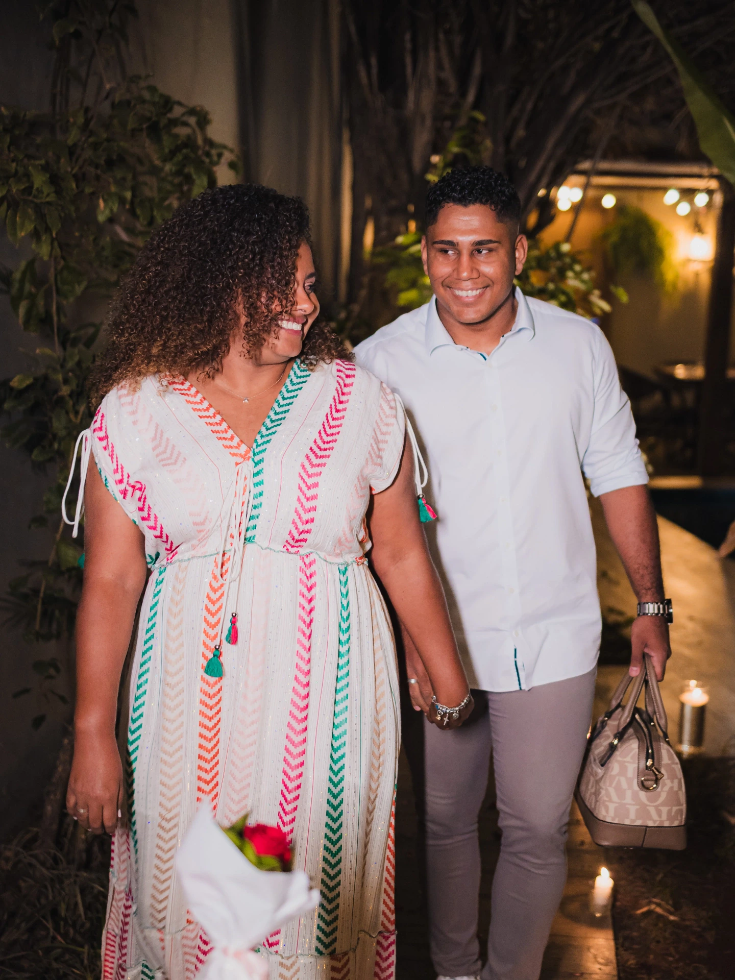 O casal sorrindo em pé no ambiente com luzes, a mulher segurando um pequeno buquê