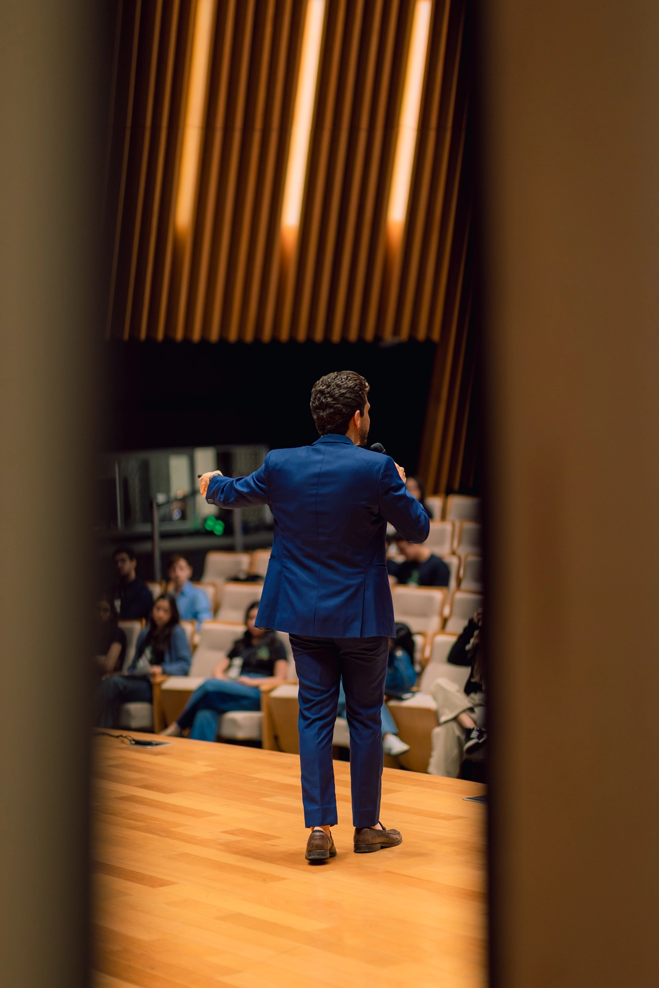 Palestrante visto de costas no palco, dirigindo-se à plateia do evento corporativo