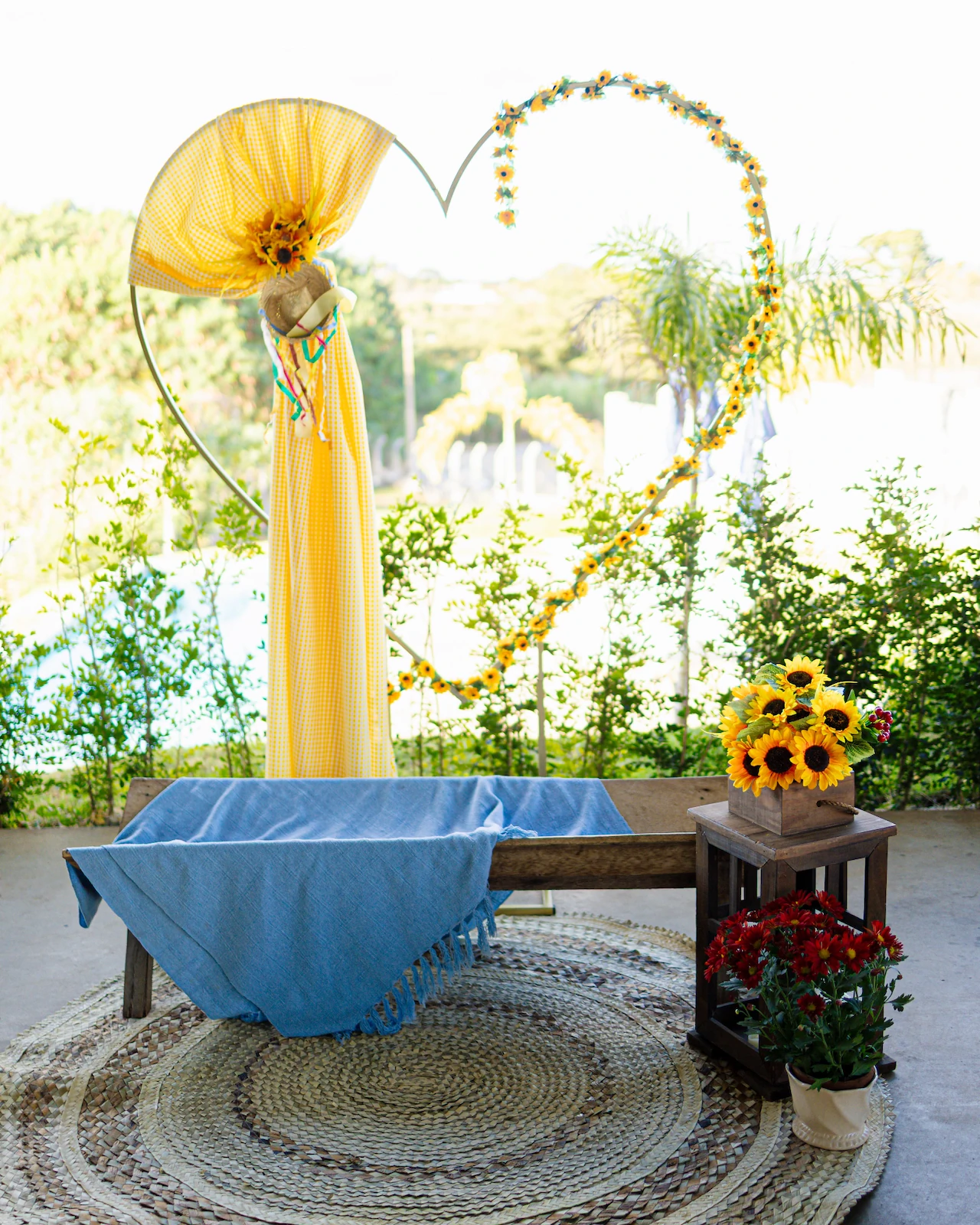 Cenário de festa junina com mesa de toalha azul, girassóis e arco em formato de coração