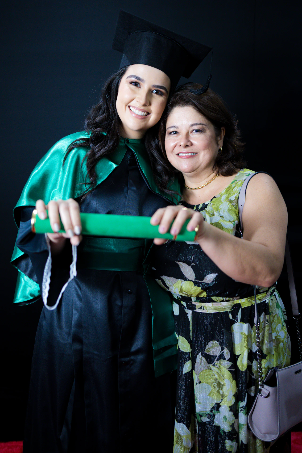 Formanda de beca abraçada com a mãe, as duas segurando o canudo