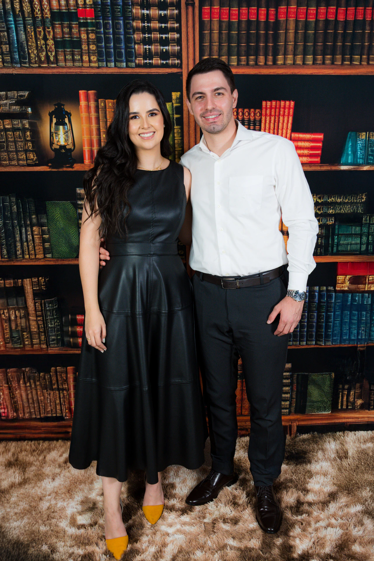 Formanda de vestido preto ao lado do namorado diante de uma estante de livros