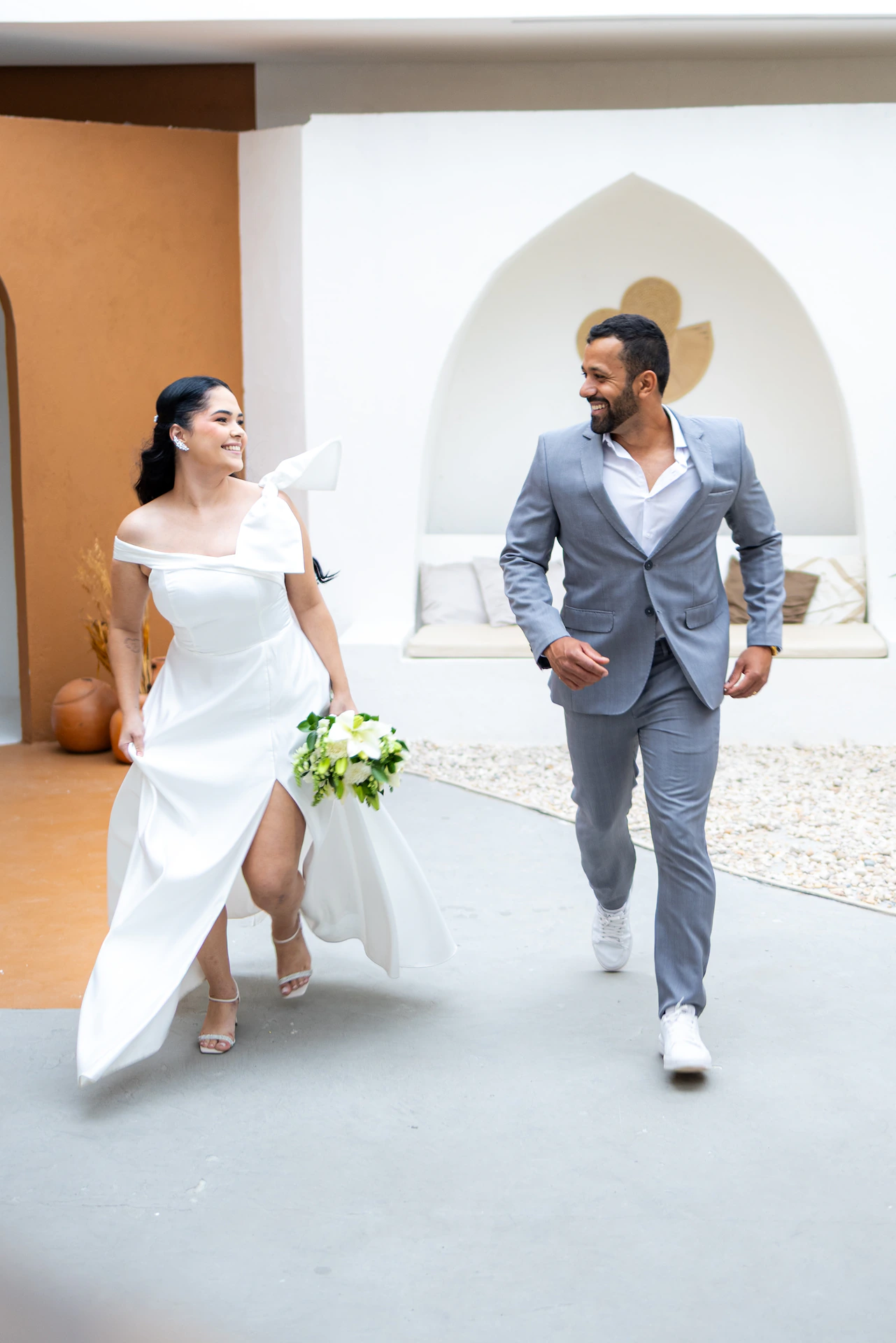 Casal caminhando e sorrindo, a noiva erguendo a barra do vestido, no ensaio pré-wedding