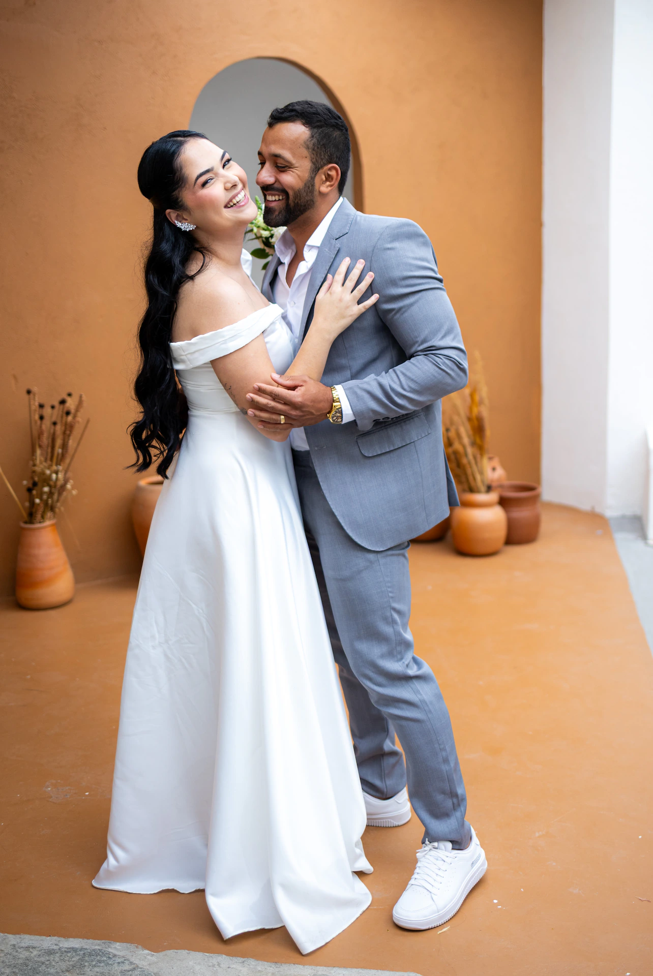 Noivo beijando a testa da noiva junto a uma parede terracota com arco