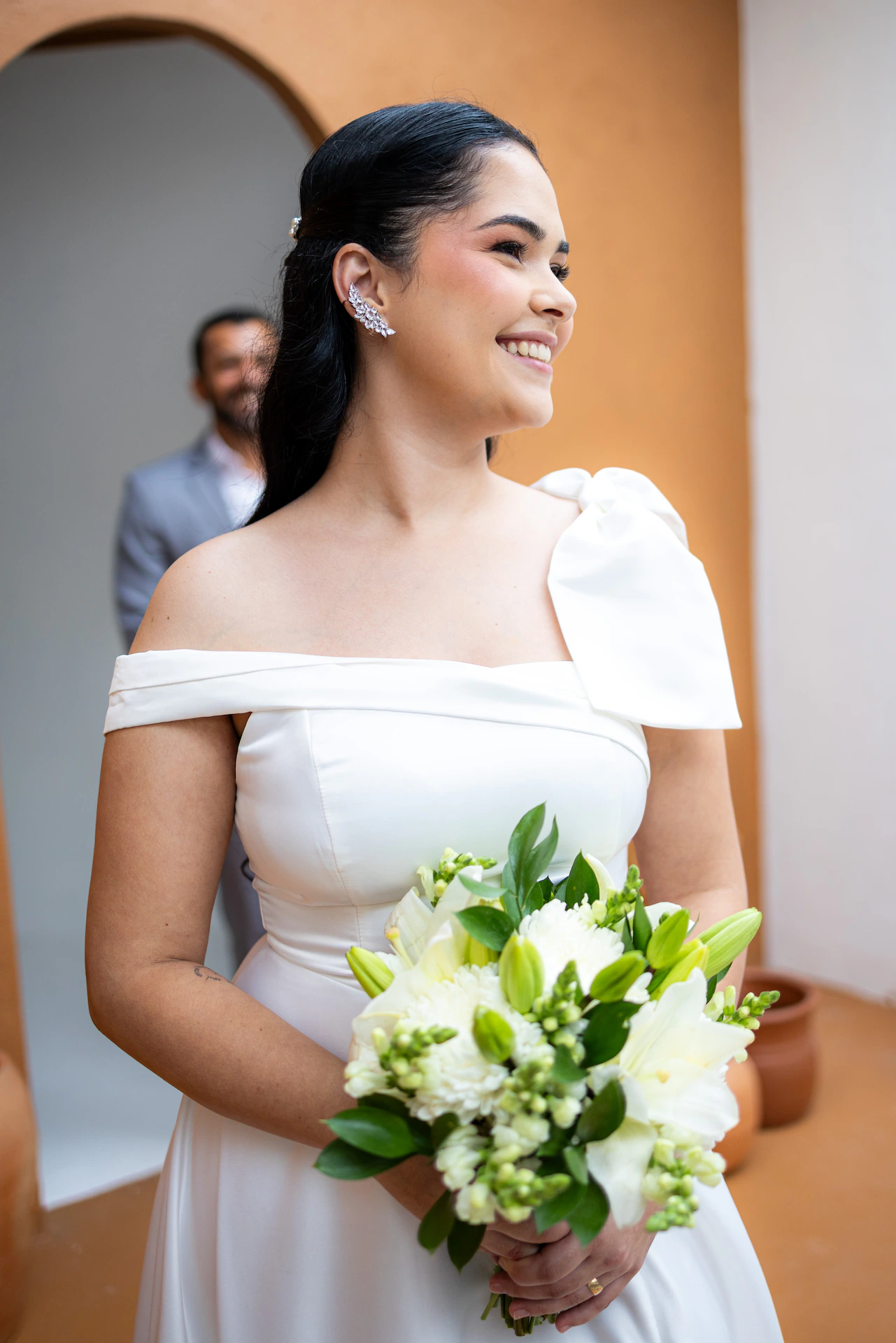 Retrato da noiva com o buquê e o noivo desfocado ao fundo, no ensaio pré-wedding